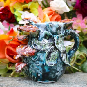 May include: A handcrafted ceramic mug with a unique design. The mug features a blue and green glaze with sculpted leaves and mushrooms in shades of brown and red. The handle is also sculpted with leaf details. The mug is set against a backdrop of colorful flowers.
