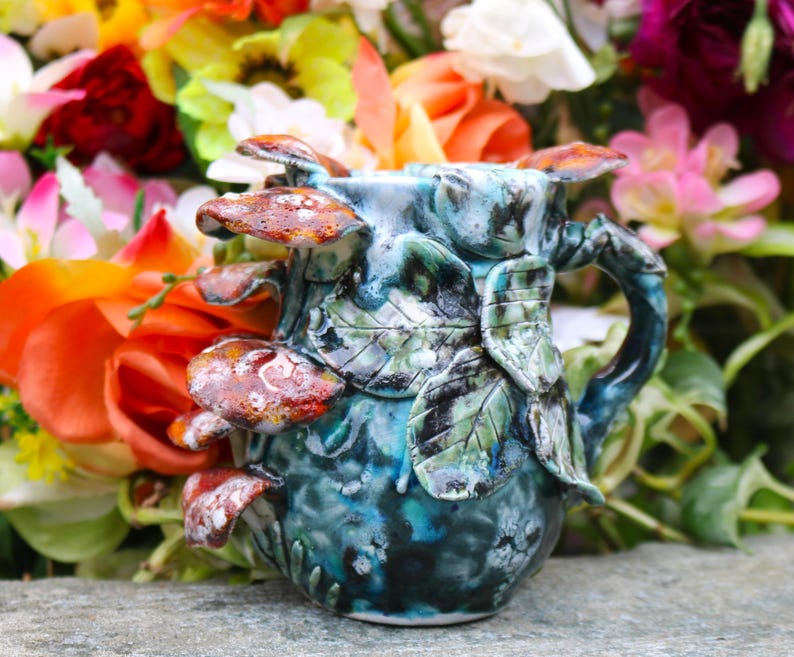 May include: A handcrafted ceramic mug with a unique design. The mug features a blue and green glaze with sculpted leaves and mushrooms in shades of brown and red. The handle is also sculpted with leaf details. The mug is set against a backdrop of colorful flowers.