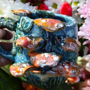 Op de afbeelding: Een blauwe keramische mok met een uniek ontwerp met een reeks overlappende, getextureerde, oranje, rode en witte paddenstoelvormige vormen.