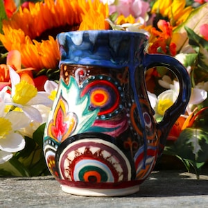 May include: A hand-painted ceramic mug with a dark brown base and a blue rim. The mug features colorful abstract designs in red, orange, green, and white. The handle is also blue. The mug is set against a backdrop of vibrant flowers.