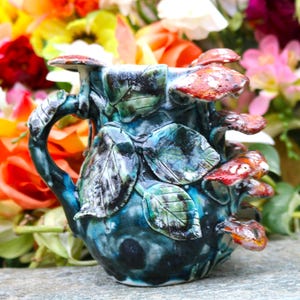 May include: A handcrafted ceramic mug with a unique mushroom and leaf design. The mug features a teal and blue glaze with red and white mushroom accents. The handle is shaped like a branch, and the leaves are green.