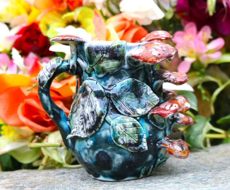 May include: A handcrafted ceramic mug with a unique mushroom and leaf design. The mug features a teal and blue glaze with red and white mushroom accents. The handle is shaped like a branch, and the leaves are green.