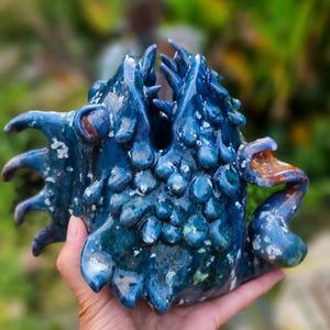 Könnte beinhalten: Eine handgeformte Keramikfigur eines Fabelwesens mit tiefblauer, glänzender Glasur, strukturierten Schuppen, ausgeprägten Flossen und zwei separaten Köpfen mit geöffneten Mäulern. Die Oberfläche zeigt gesprenkelte weiße und blaue Farbtöne.