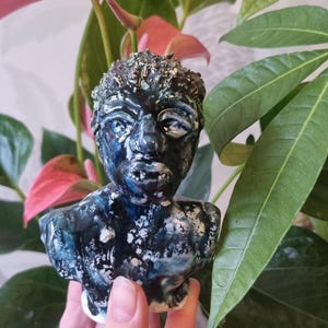 以下が含まれることがあります： 濃い青と白の釉薬を施した陶器の胸像。彫刻は、詳細な特徴と質感のある髪を持つ人間の顔を描いています。胸像は、緑とピンクの葉の前に保持されています。