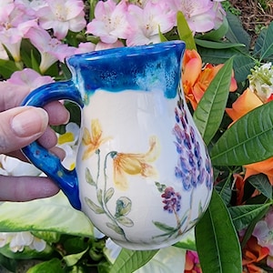 以下が含まれることがあります： 青い縁取りと花柄の白い陶器のマグカップ。マグカップには持ち手があり、黄色、紫、緑の花が描かれています。