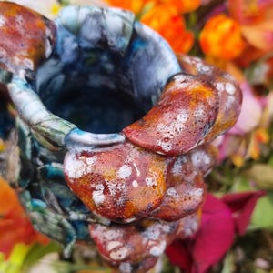 May include: A handcrafted ceramic vase with a unique, textured design. The vase features a blend of colors, including shades of blue, brown, and white. The exterior has a layered, organic appearance, with a rough, uneven surface. The vase is surrounded by colorful flowers.