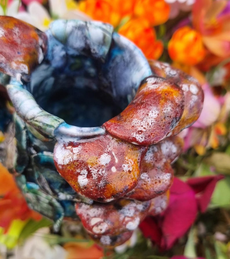 May include: A handcrafted ceramic vase with a unique, textured design. The vase features a blend of colors, including shades of blue, brown, and white. The exterior has a layered, organic appearance, with a rough, uneven surface. The vase is surrounded by colorful flowers.