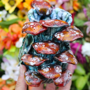 May include: A handcrafted ceramic mushroom sculpture with a cluster of reddish-brown caps speckled with white. The base is a textured teal color. The sculpture is held in a hand against a blurred background of colorful flowers.
