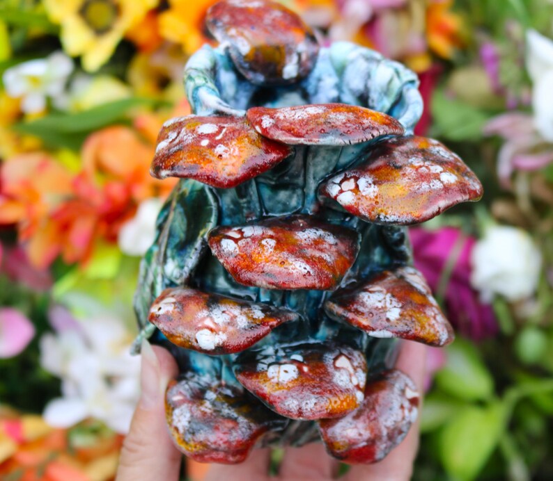 May include: A handcrafted ceramic mushroom sculpture with a cluster of reddish-brown caps speckled with white. The base is a textured teal color. The sculpture is held in a hand against a blurred background of colorful flowers.