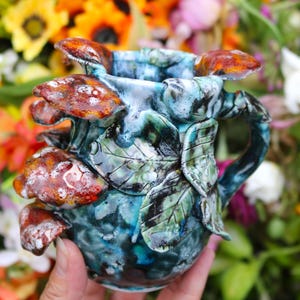 May include: A ceramic mug with a unique design featuring brown and white mushroom accents. The mug has a blue and green mottled glaze, with leaf and vine details. The handle is integrated into the design. The mug is held in a hand, with a blurred floral background.