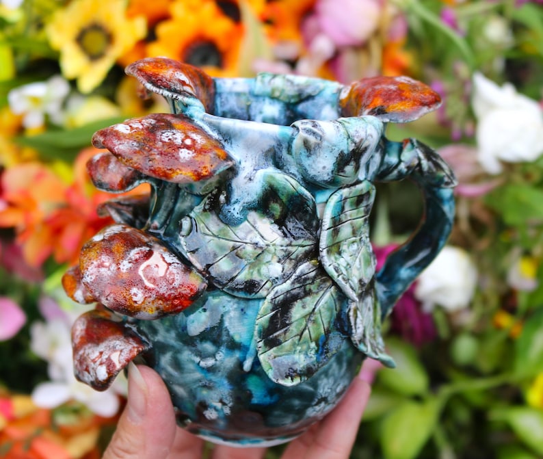 May include: A ceramic mug with a unique design featuring brown and white mushroom accents. The mug has a blue and green mottled glaze, with leaf and vine details. The handle is integrated into the design. The mug is held in a hand, with a blurred floral background.