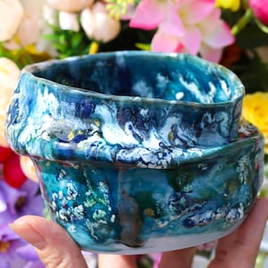 May include: A handmade ceramic bowl with a blue, green and white marbled glaze. The bowl has a unique, irregular shape and a glossy finish.
