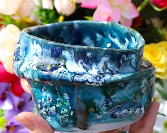 May include: A handmade ceramic bowl with a blue, green, and white marbled glaze. The bowl has a unique, irregular shape and a glossy finish.