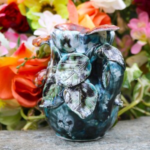 May include: A handcrafted ceramic pitcher with a leaf design. The pitcher is primarily teal and blue, with accents of green, brown, and white. The handle and body are adorned with sculpted leaves. The pitcher is approximately 6 inches tall.