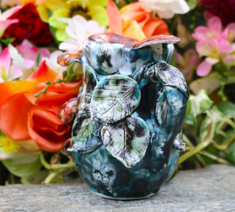 May include: A handcrafted ceramic pitcher with a leaf design. The pitcher is primarily teal and blue, with accents of green, brown, and white. The handle and body are adorned with sculpted leaves. The pitcher is approximately 6 inches tall.