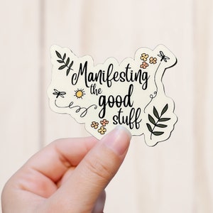 May include: A cream-colored sticker with the phrase "Manifesting the good stuff" in black script. The sticker features small illustrations of a sun, flowers, dragonflies, and leaves. The sticker is held in a hand.