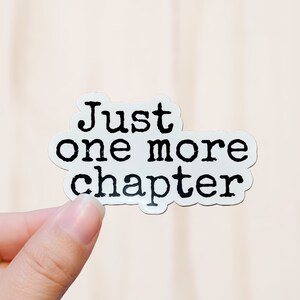 May include: White sticker with the text "Just one more chapter" in black, bold, typewriter-style font. The sticker has a rounded, irregular shape and is held against a light beige background.