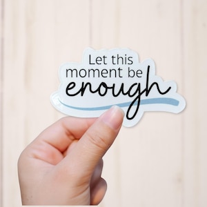 May include: A white die-cut sticker featuring the black text Let this moment be enough with a light blue wavy line underneath. The sticker has a smooth, irregular shape and is held against a light, neutral-toned background.