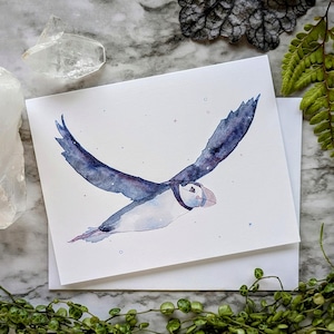 May include: A rectangular greeting card featuring a watercolour illustration of a puffin in flight. The bird has dark blue wings and a white body with a distinctively coloured beak. The background of the illustration is white with small scattered dots.