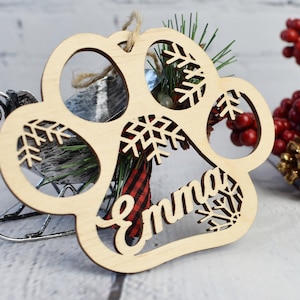 May include: A wooden paw print Christmas bauble with a snowflake design and the name "Emma" cut out. The bauble is decorated with a red and black checked ribbon and a sprig of pine.