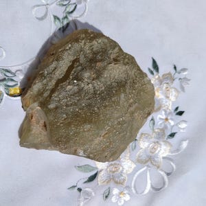 Puede incluir: Un trozo grande de vidrio verde y marrón de forma irregular con una superficie rugosa y texturizada. El vidrio es translúcido y parece haberse formado naturalmente.