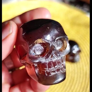 May include: A carved, translucent skull figurine with a dark amber hue. The skull features detailed facial features, including eye sockets and a defined jawline. A second, smaller skull is visible in the background.