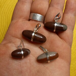 Op de afbeelding: Vier bruine en grijze ovale hangers met zilveren accenten. De hangers liggen op een hand.
