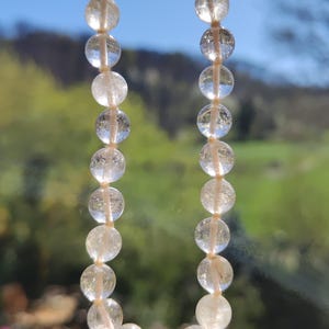 May include: A strand of clear quartz beads with a subtle golden shimmer. The beads are strung on a white cord and are arranged in a circular shape.