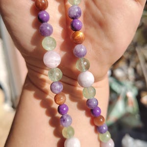 Peut inclure: Un collier de perles composé de pierres précieuses colorées. Les perles rondes sont de couleurs violettes, vertes, marron et blanches. Le collier est tenu dans une main, les perles étant enfilées ensemble.