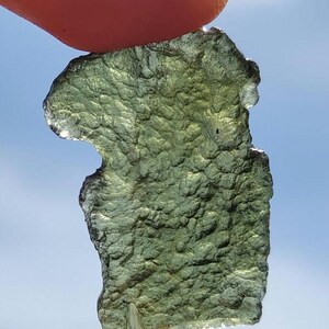 Könnte beinhalten: Ein grünes, durchsichtiges, unregelmäßig geformtes Stück Moldavit, eine Art Tektit, das in einer Hand vor blauem Himmel gehalten wird.