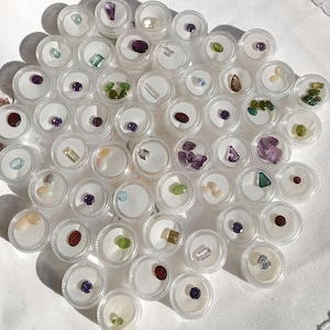May include: A collection of small, clear plastic containers, each holding a different gemstone. The gemstones vary in color, including purple, green, blue, and clear. The containers are arranged on a white surface, creating a display of various precious stones.