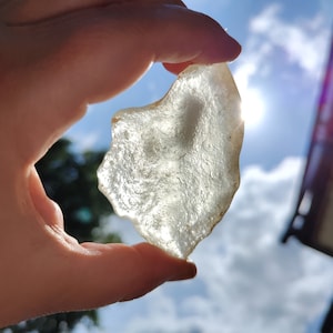May include: A clear, translucent piece of sea glass, with a rough, irregular edge, held in a hand against a bright blue sky with white clouds.