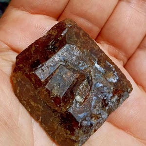 Op de afbeelding: Een gefacetteerd, donkerbruin mineraal specimen met een kristalstructuur. De steen heeft een vierkante vorm met meerdere facetten die licht reflecteren. Het oppervlak heeft een mix van bruine en witte vlekken. Uniek.