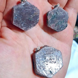 Op de afbeelding: Drie zeshoekige hangers van ruwe, gefacetteerde edelstenen in paars en zilver. De hangers hebben een zilveren oogje. De edelstenen hebben een ruwe, natuurlijke textuur. De hangers worden in een hand gehouden.