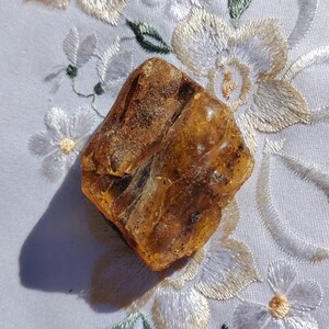 May include: A rough, amber-colored gemstone with a natural, irregular shape. The stone has a translucent quality, allowing light to pass through. It rests on a white fabric with embroidered floral patterns in white, cream, and green.