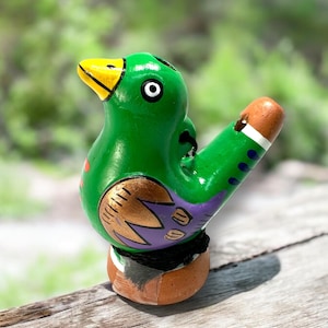 May include: A green ceramic bird whistle with purple, gold, and white accents. The bird has a yellow beak and a white eye.