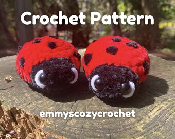 May include: Two plush, crocheted ladybugs with red bodies, black spots, and black heads with white-rimmed eyes. The text "Crochet Pattern" is above the ladybugs, and "emmyscozycrochet" is below. The ladybugs are on a wooden surface.