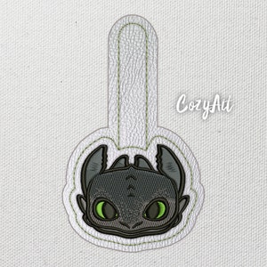 Könnte beinhalten: Ein gestickter Aufnäher mit dem Gesicht eines Drachen, mit schwarzen und grünen Nähten auf weißem Hintergrund. Der Aufnäher ist an einer weißen Stoffschlaufe befestigt.