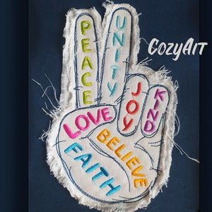 Könnte beinhalten: Ein weißes Stoff-Friedenszeichen mit den Wörtern "Peace", "Love", "Faith", "Believe", "Joy", "Unity" und "Kind", die in verschiedenen Farben gestickt sind.