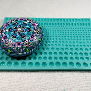Peut inclure: Une pierre ronde peinte à la main avec un motif mandala détaillé en turquoise, rose et violet, accentué de points argentés et d'une perle centrale. Elle repose sur un moule en silicone turquoise avec des empreintes circulaires de différentes tailles.