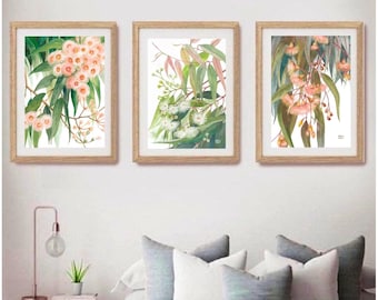 May include: Three framed botanical prints featuring detailed illustrations of eucalyptus branches with pink and white flowers. The prints are displayed on a white wall above a bed with gray and white pillows.