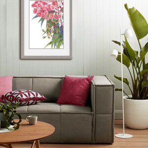 May include: A framed print of a watercolour painting of pink flowers with green leaves. The print is hanging on a white wall above a grey sofa with red and white cushions.