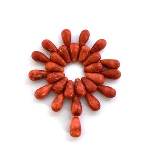 May include: A collection of teardrop-shaped, reddish-orange coral beads arranged in a circular pattern. The beads are smooth and polished, with variations in color and texture. The arrangement is centered on a white background.