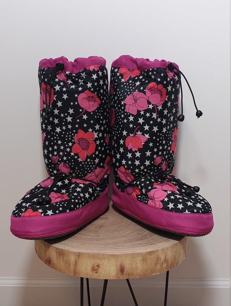 May include: A pair of black and pink fleece boot covers with a floral and star pattern. The boot covers have a drawstring at the top and a pink trim around the bottom.