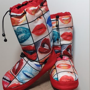 May include: A pair of red and white slipper boots with a print of colorful lips. The boots have a drawstring at the top and a red trim around the bottom.