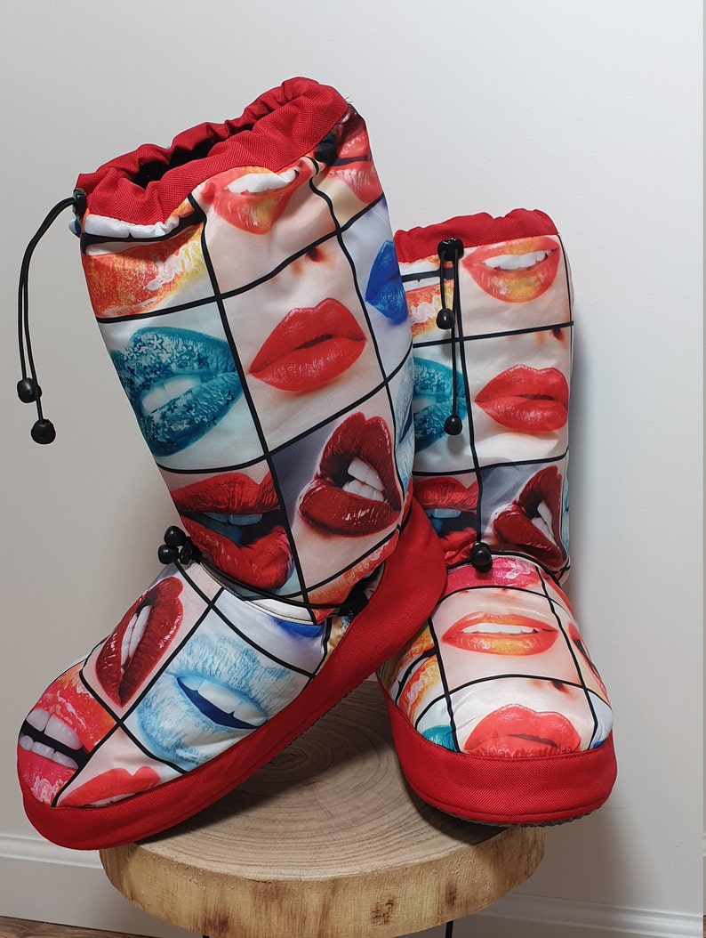 May include: A pair of red and white slipper boots with a print of colourful lips. The boots have a drawstring at the top and a red trim around the bottom.