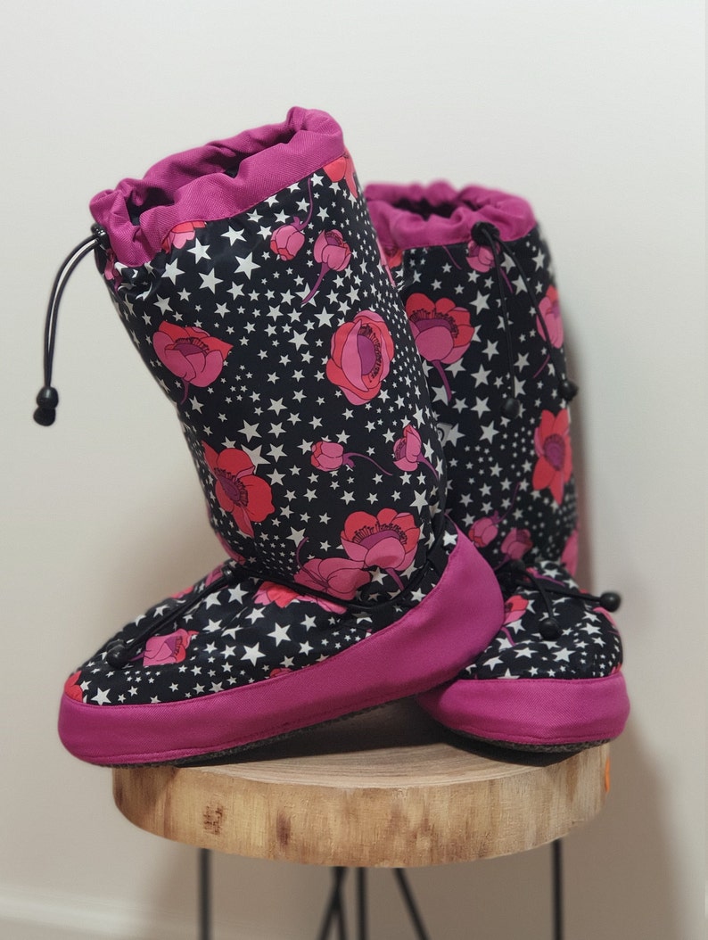 May include: A pair of pink and black fleece lined indoor boots with a drawstring closure. The boots have a floral and star pattern.