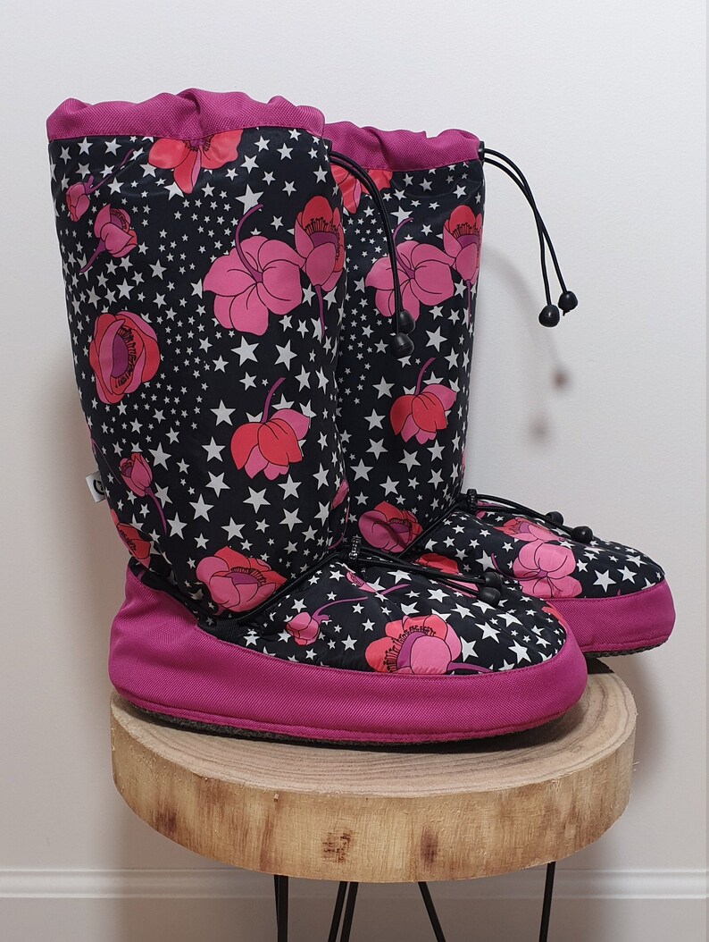 May include: A pair of pink and black fleece boot liners with a floral and star pattern. The boot liners have a drawstring closure at the top.