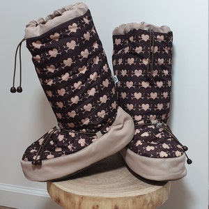 May include: A pair of brown and beige fleece boot covers with a pink heart pattern. The boot covers have a drawstring at the top and a brown cord.