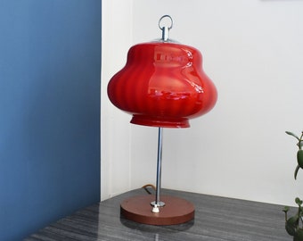 May include: A striking red table lamp featuring a rounded shade, chrome stem, and a circular wooden base. The lamp sits on a grey table. The background includes a blue wall and a decorative clock. The lamp's height is approximately 30 cm.